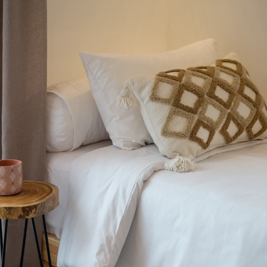 Close-up of bed with white linens and textured boho cushion beside a wooden side table